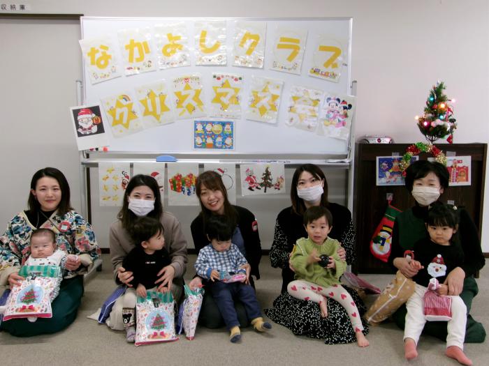 クリスマス会の看板の前で参加した親子たちが貰ったプレゼントを持って横一列に座っている様子