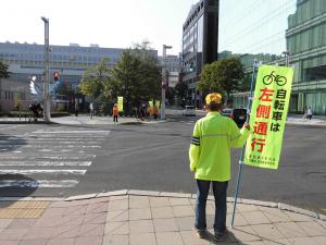 交差点で「自転車は左側通行」の旗を持ち、安全運転を呼び掛けている様子