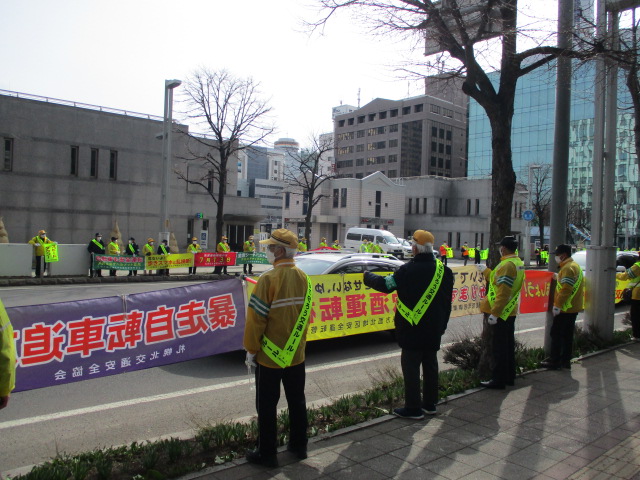 参加者が車道の両側で交通安全標語が記された横断幕を掲げ、啓発活動をしている様子