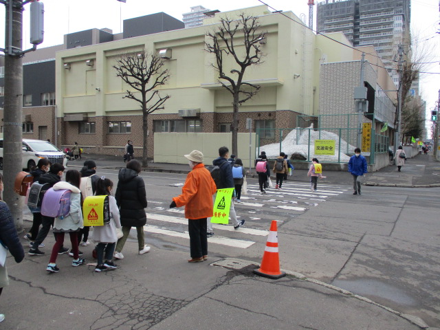 横断歩道を渡って登校する小学生を、地域の方が「横断中」の旗を持って誘導している様子