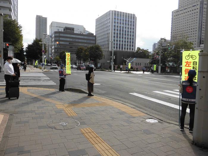 歩道に自転車走行安全運転を呼びかける旗を持って立つ交通安全指導員たちの様子
