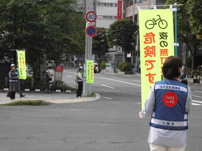 交差点で自転車走行安全運転を呼びかける旗を持って立つ交通安全指導員の様子