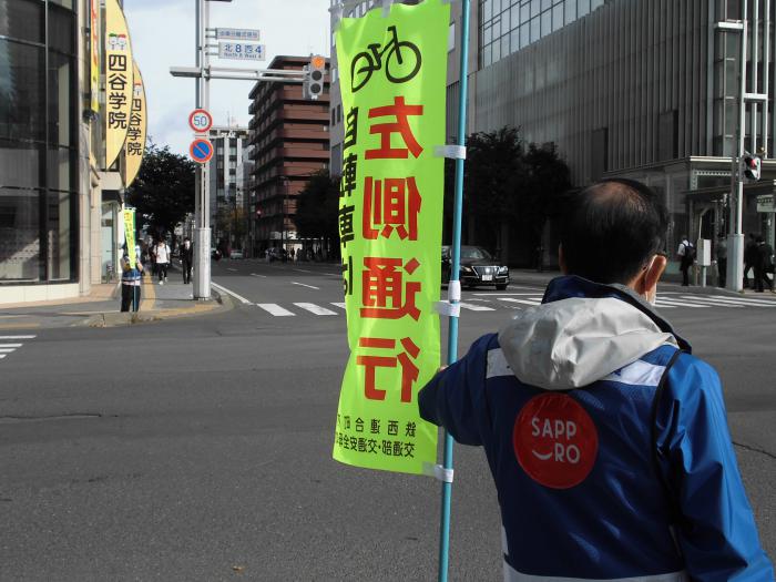 交差点で自転車走行安全運転を呼びかける旗を持って立つ交通安全指導員の様子