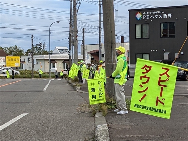 「スピードダウン」「シートベルト着用」と書かれた旗を掲げて沿道に並ぶ参加者