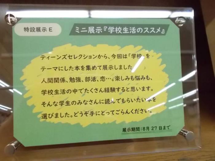 展示の看板です。アクリルフレームにハガキ程度のサイズで掲出しています。薄緑色の背景に黄色い絵の具で塗りつけたようなデザインです。塗り付けた箇所には次のメッセージを添えています。ティーンズセレクションから、今回は「学校」をテーマにした本を集めて展示しました。人間関係、勉強、部活、恋…。楽しみも悩みも、学校生活の中でたくさん経験すると思います。そんな学生のみなさんに読んでもらいたい本を選びました。どうぞ手にとってごらんください。