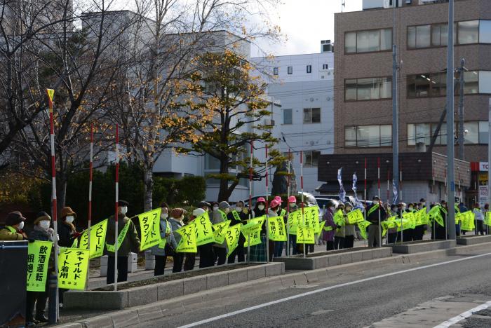 スピードダウンなどとメッセージが書かれた黄色い旗を振りながら通行する車両に訴えかける様子