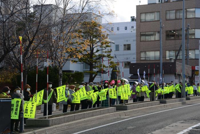 スピードダウンなどとメッセージが書かれた黄色い旗を振りながら通行する車両に訴えかける様子